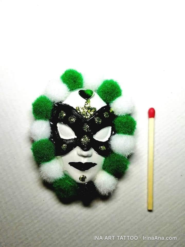 A martisor shaped like a white and black Venetian mask, surrounded by green and white pom-poms. A matchstick is beside it.
