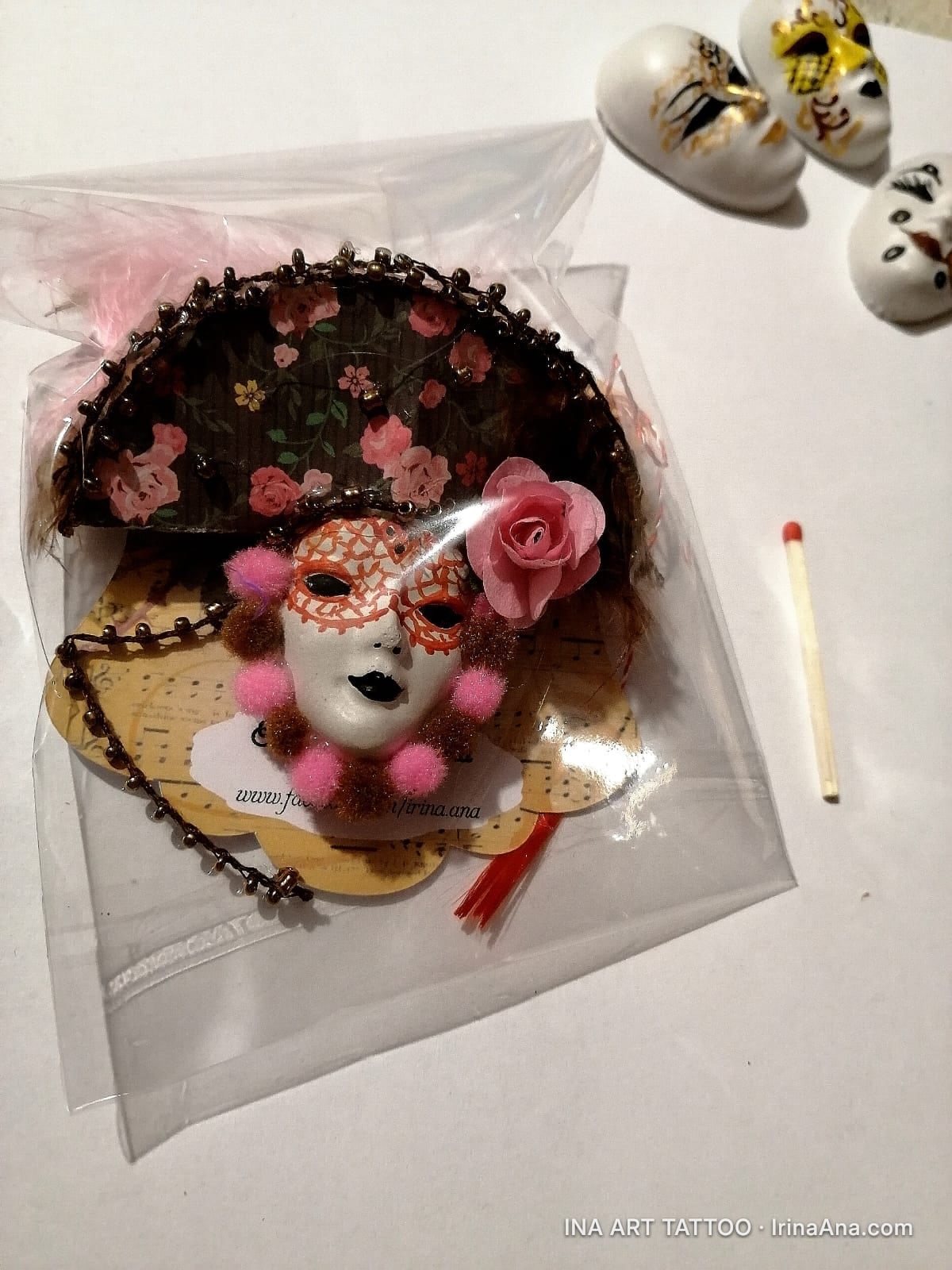 Handmade Mărțișor in the shape of a Venetian mask, with a pink rose and feathers, packaged in clear plastic. A matchstick an…