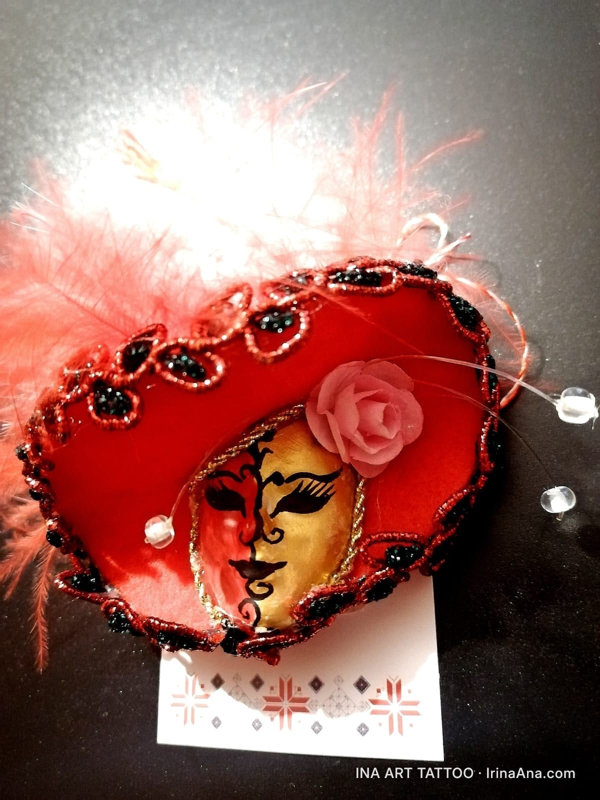 Mărțișor in the shape of a red Venetian mask with feathers, decorated with a pink rose and black beads.
