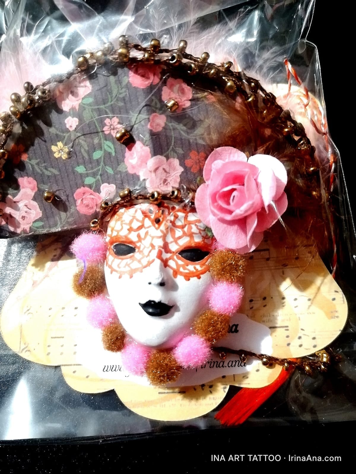 Handmade Mărțișor with a white Venetian mask, red details, surrounded by pink and brown pompoms, on a floral background.
