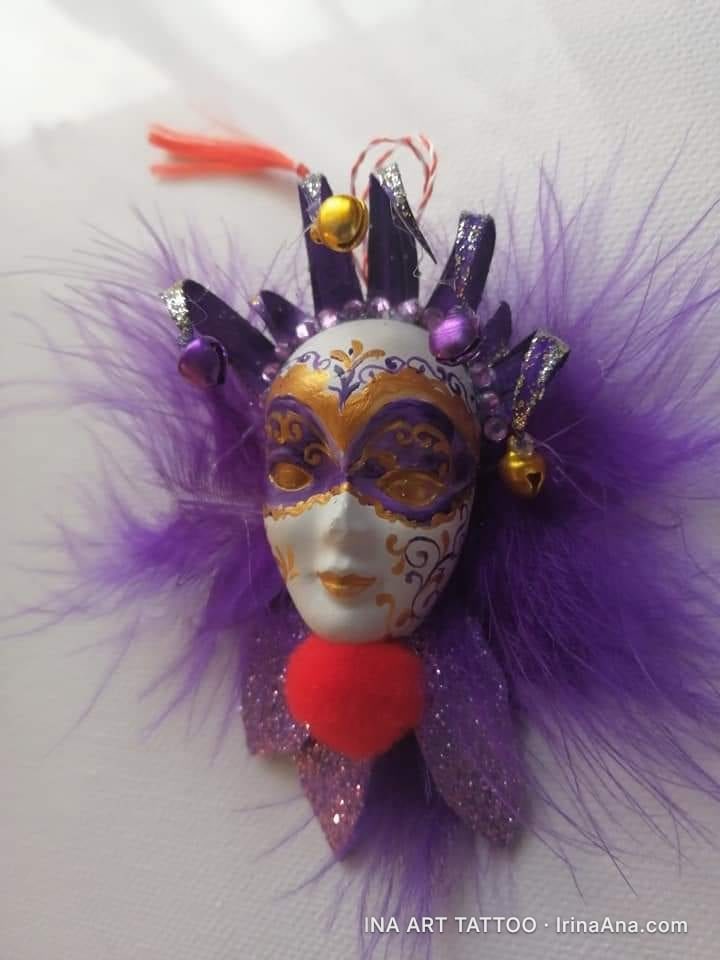 Mărțișor in the shape of a white Venetian mask, with gold and purple details, surrounded by purple feathers and a red pompom.