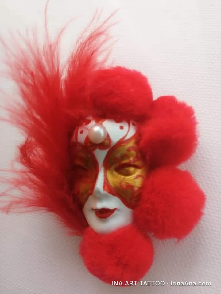 A white, gold, and red Venetian mask, decorated with red feathers and pom-poms, with a pearl on its forehead.