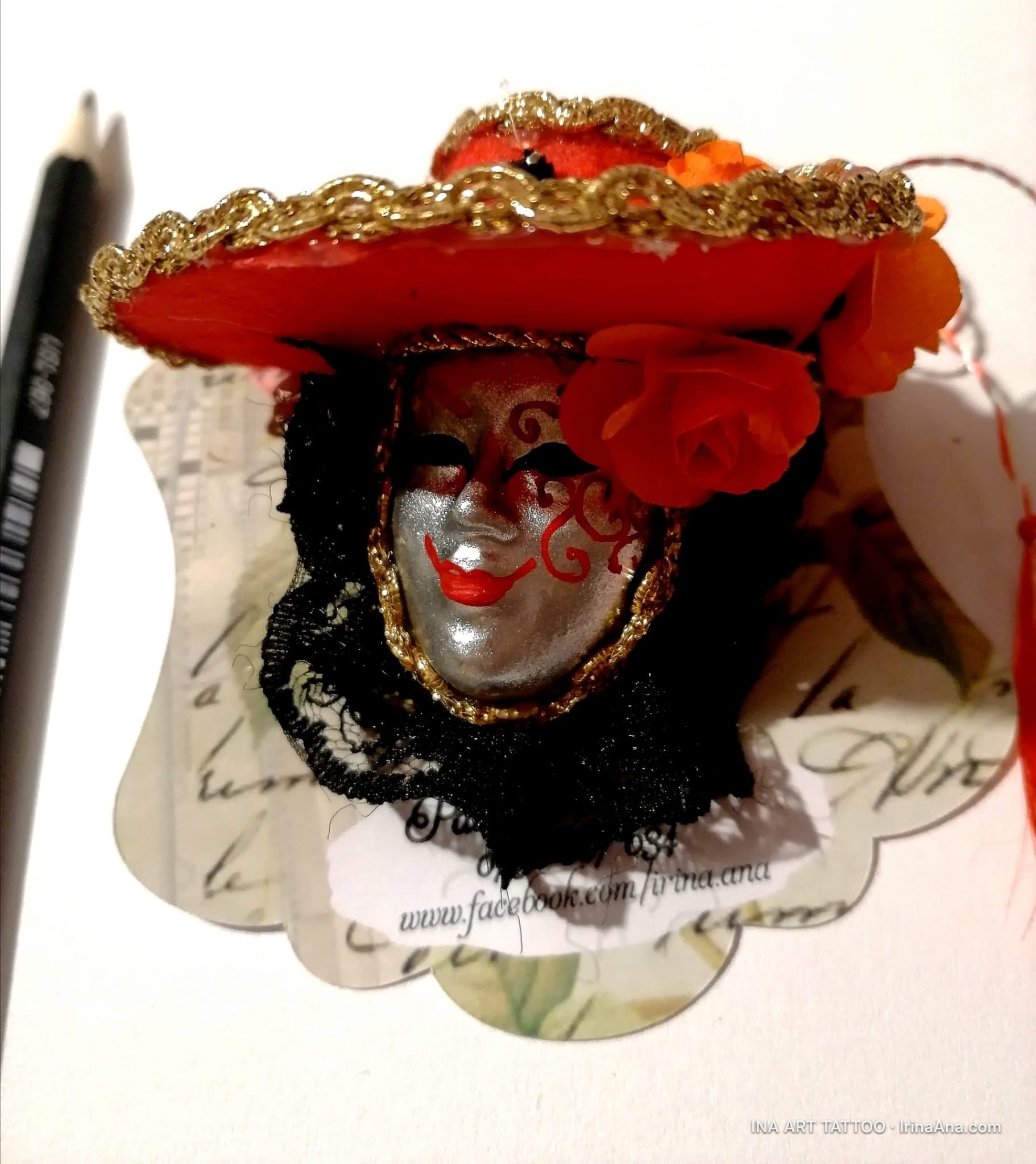 A martisor: silver Venetian mask with red lips, wearing a red hat with gold trim and orange flowers.