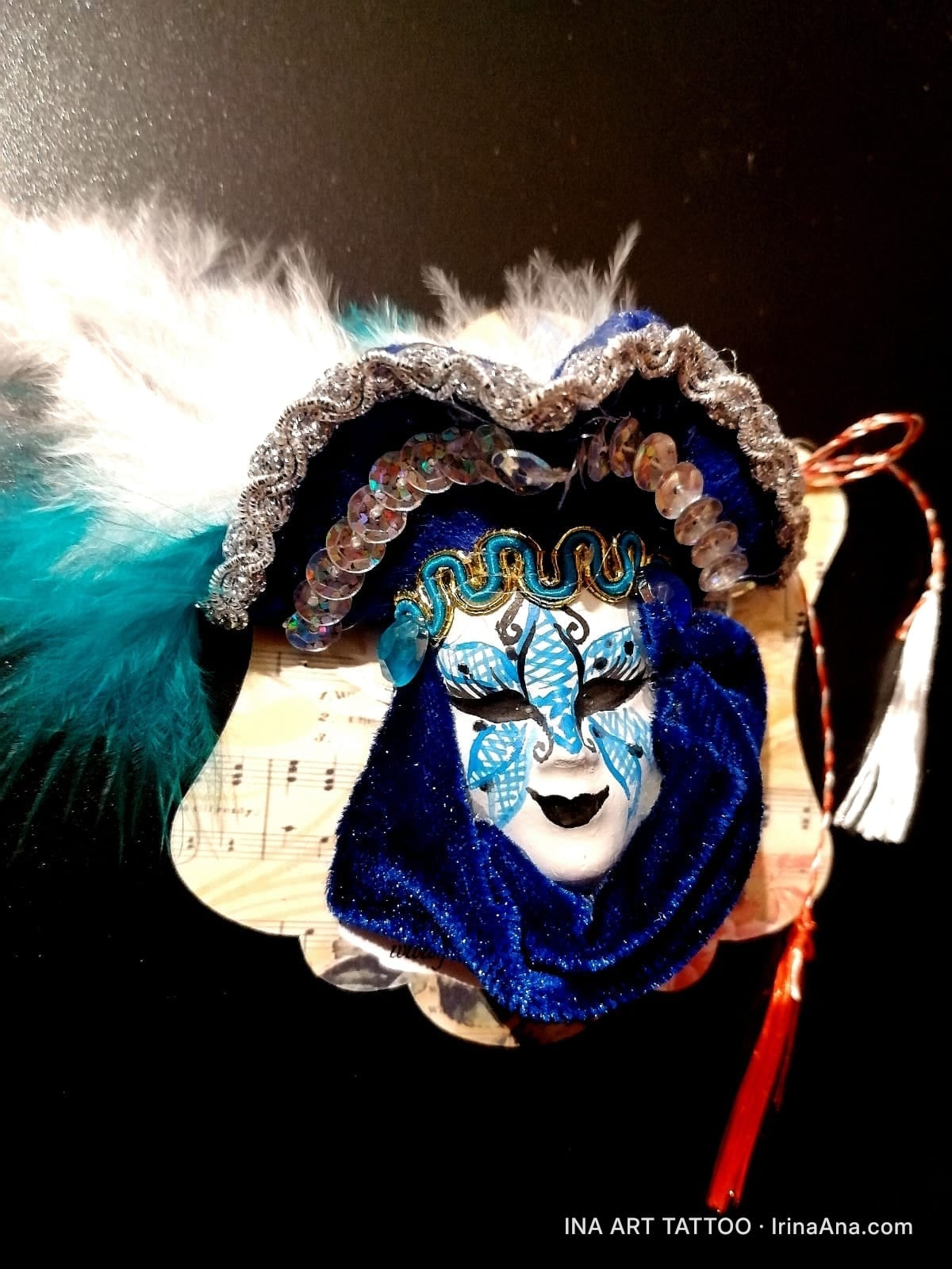 Mărțișor with a white and blue Venetian mask, with white and turquoise feathers, on a black background.