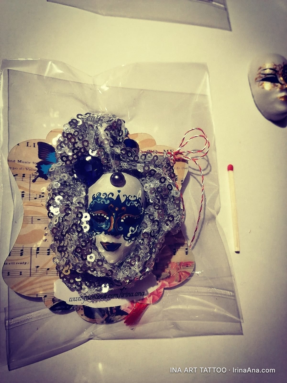 Martisor in the shape of a white Venetian mask with blue details, surrounded by silver sequins and a red bow.