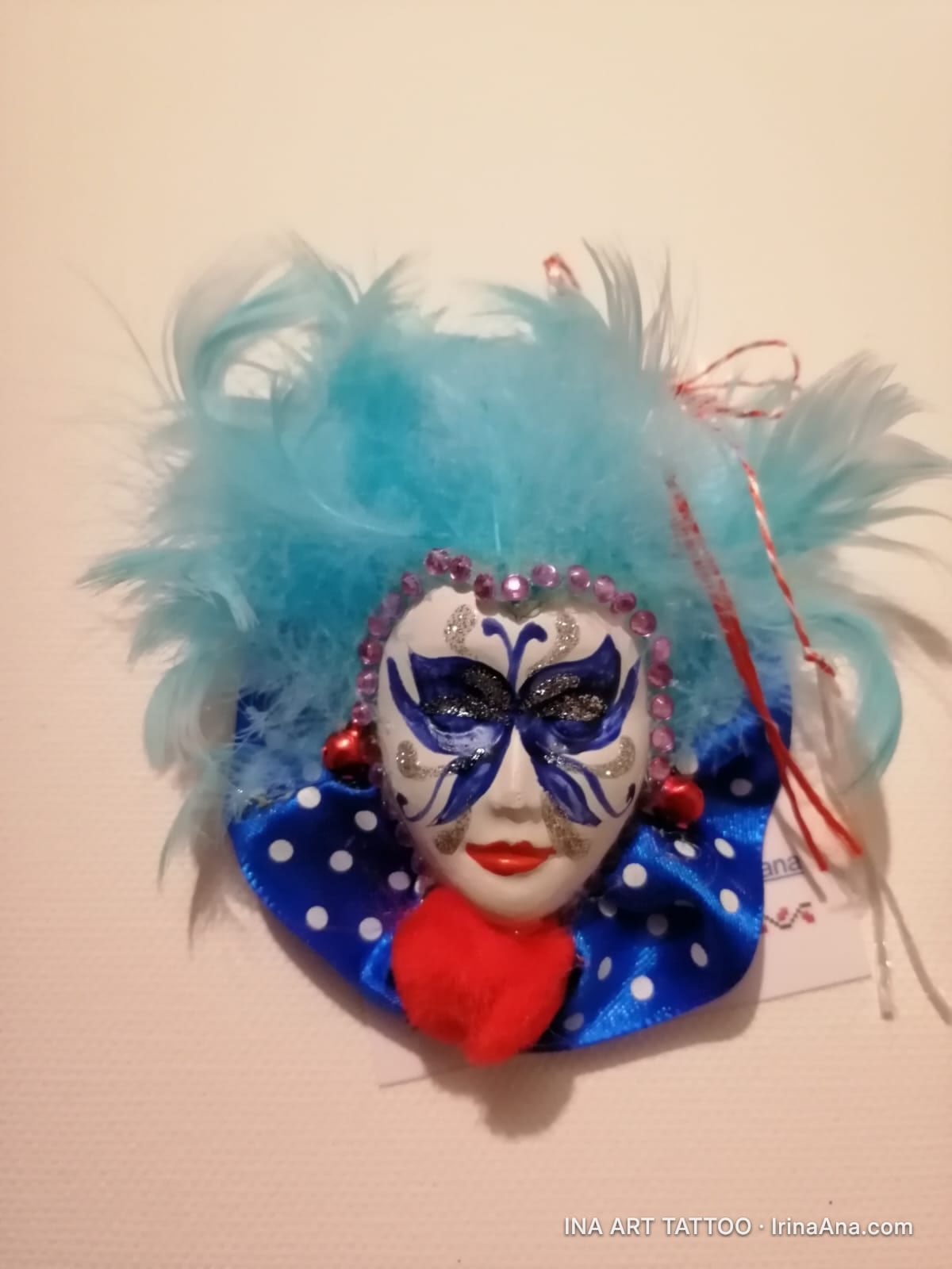 Mărțișor in the shape of a white Venetian mask with a blue mask, turquoise feathers, a blue polka-dot collar, and a red hear…