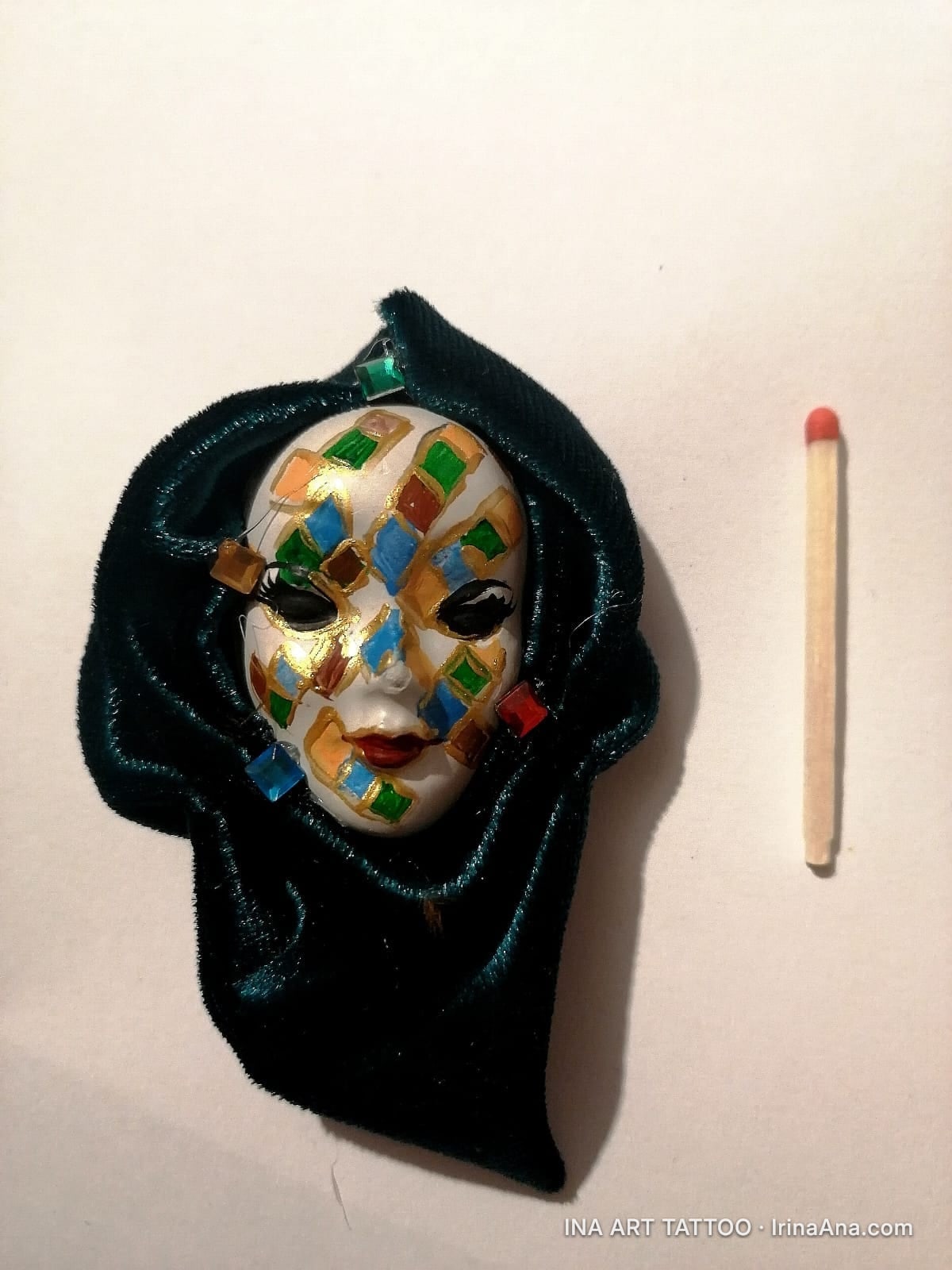 Handmade brooch of a white Venetian mask, decorated with colorful diamonds, wrapped in dark green velvet. A matchstick is ne…