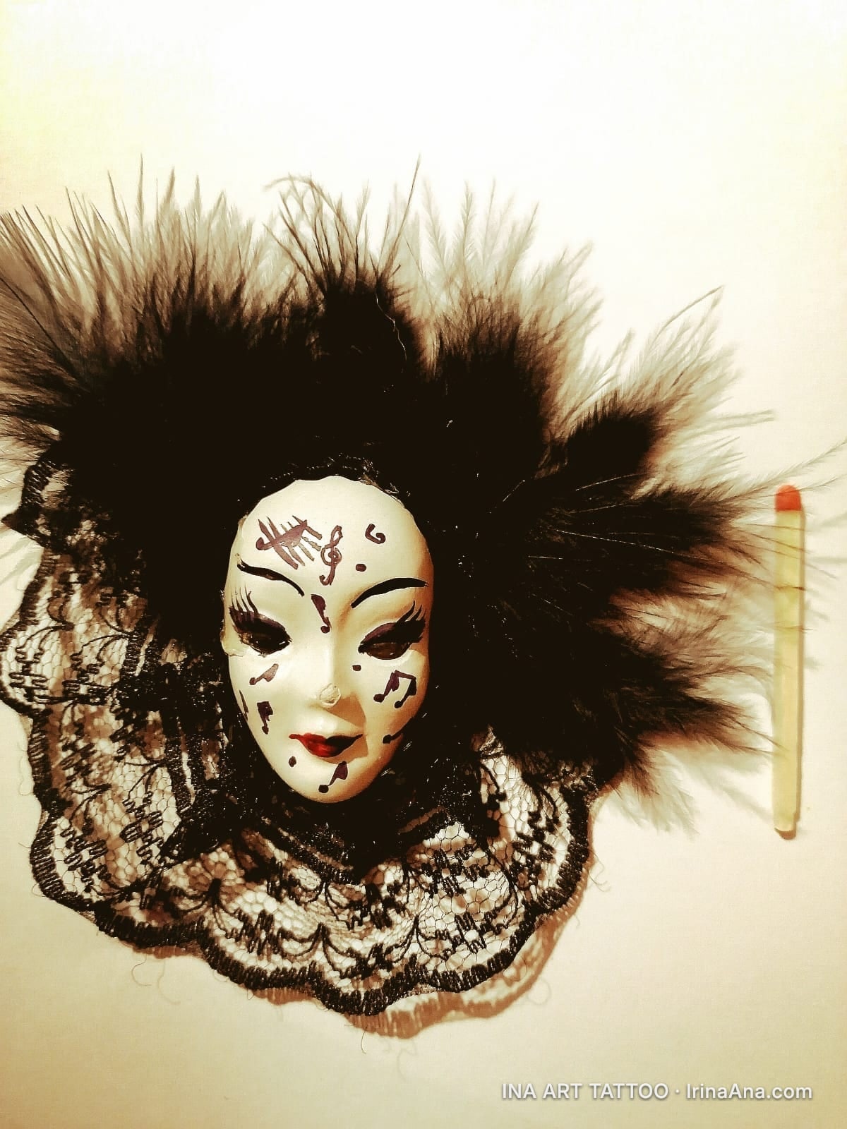 A white Venetian mask with black musical notes, surrounded by black feathers and black lace, on a light background.