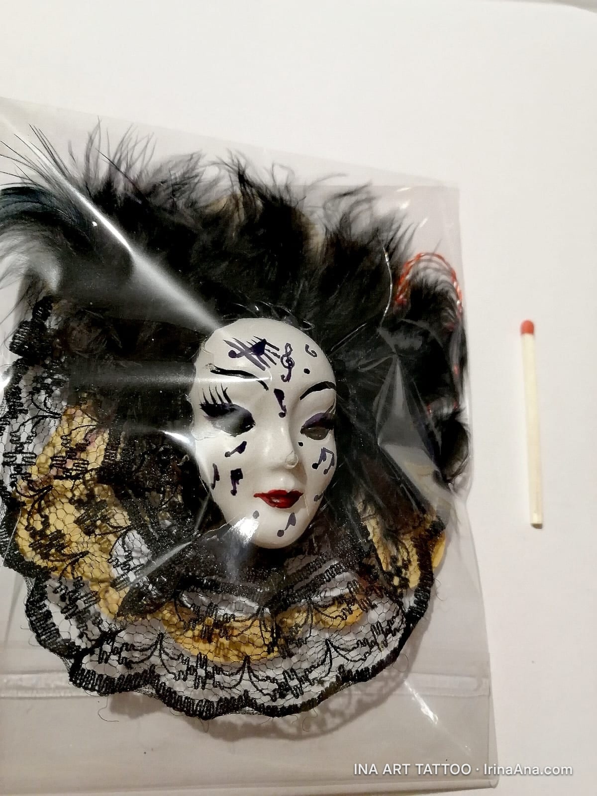 A white Venetian mask with black musical notes, surrounded by black feathers and black and gold lace, in a plastic bag.