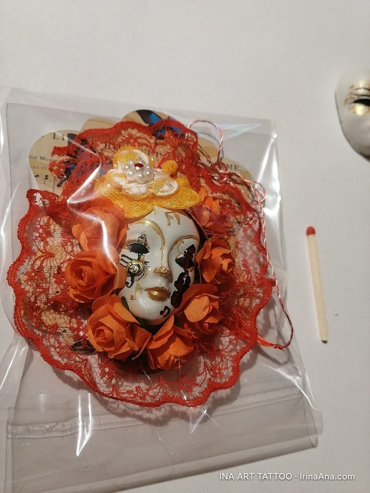 Mărțișor: white Venetian mask with gold and black details, surrounded by orange roses and red lace.