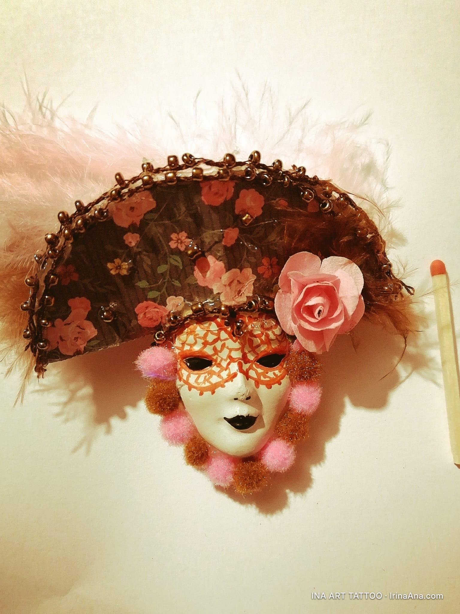 A Venetian mask decorated with pink feathers, a pink flower, brown beads, and pink and brown tassels, on a white background.