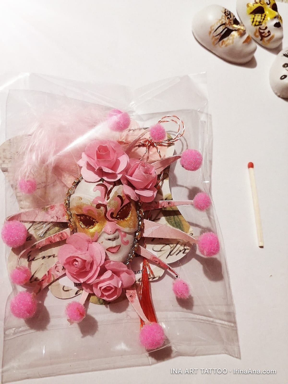 A Venetian mask decorated with pink roses, feathers, and pom-poms, packaged in a clear bag. A matchstick is next to it.