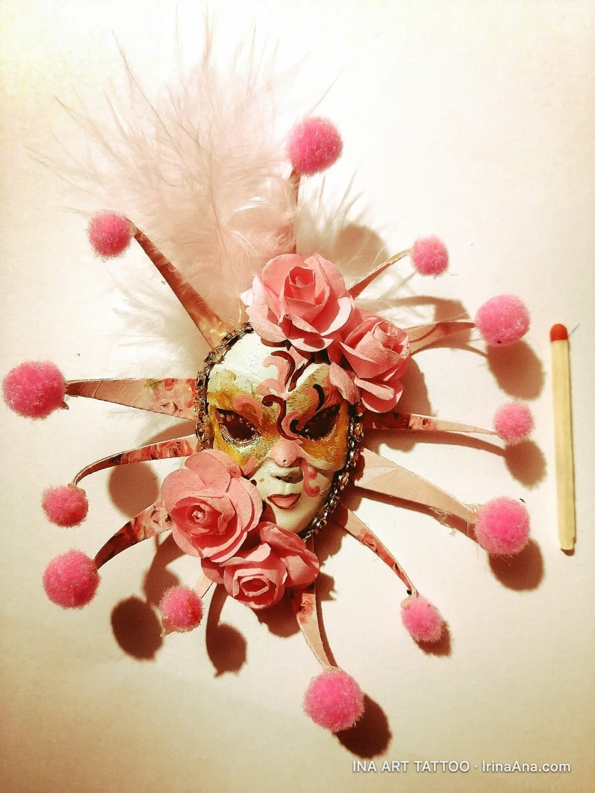 A Venetian mask decorated with pink roses, feathers, and fluffy pompoms, next to a matchstick.