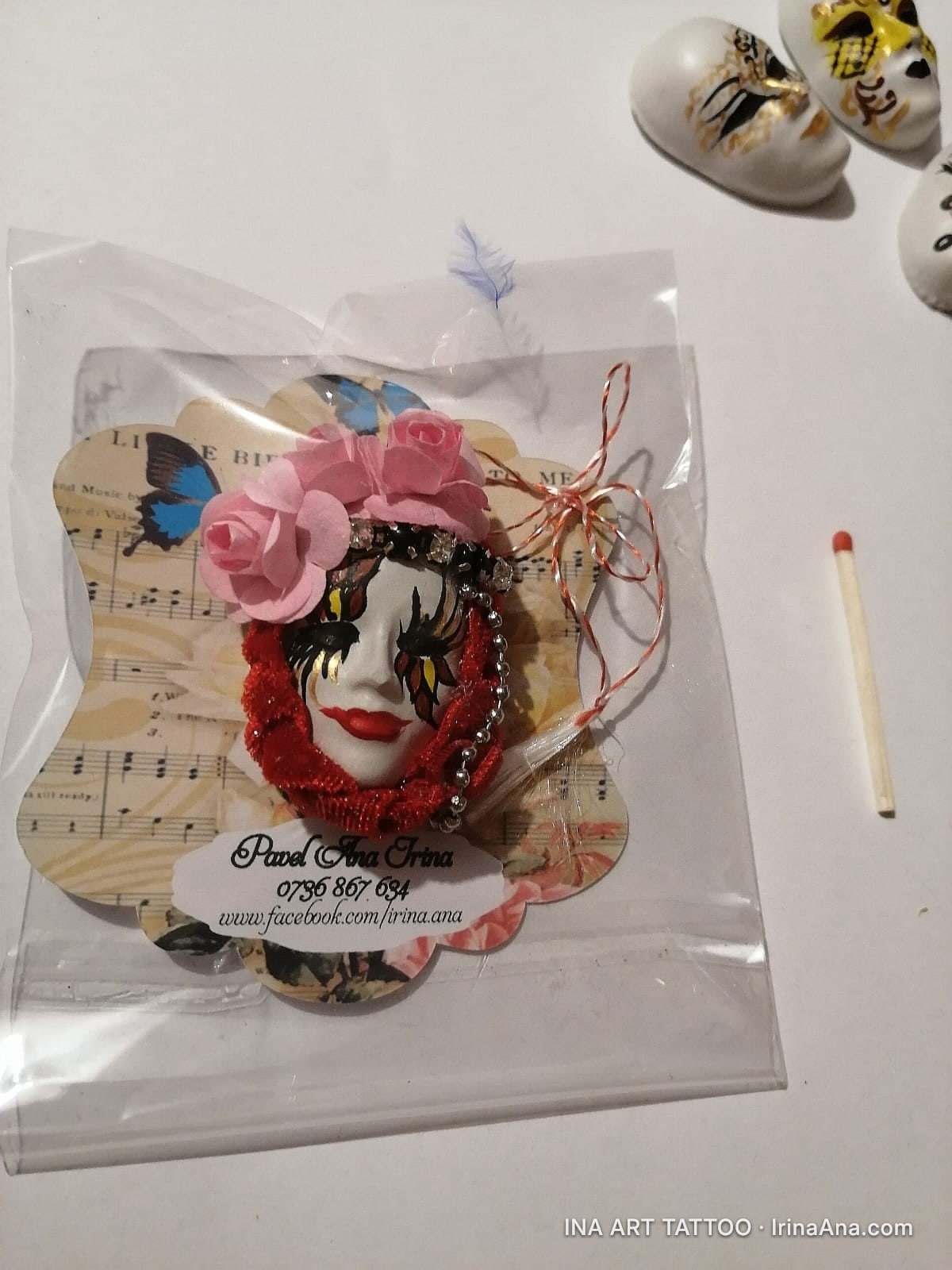 A handmade mărțișor featuring a white Venetian mask with gold and black makeup, framed by pink roses and a red scarf, on a m…