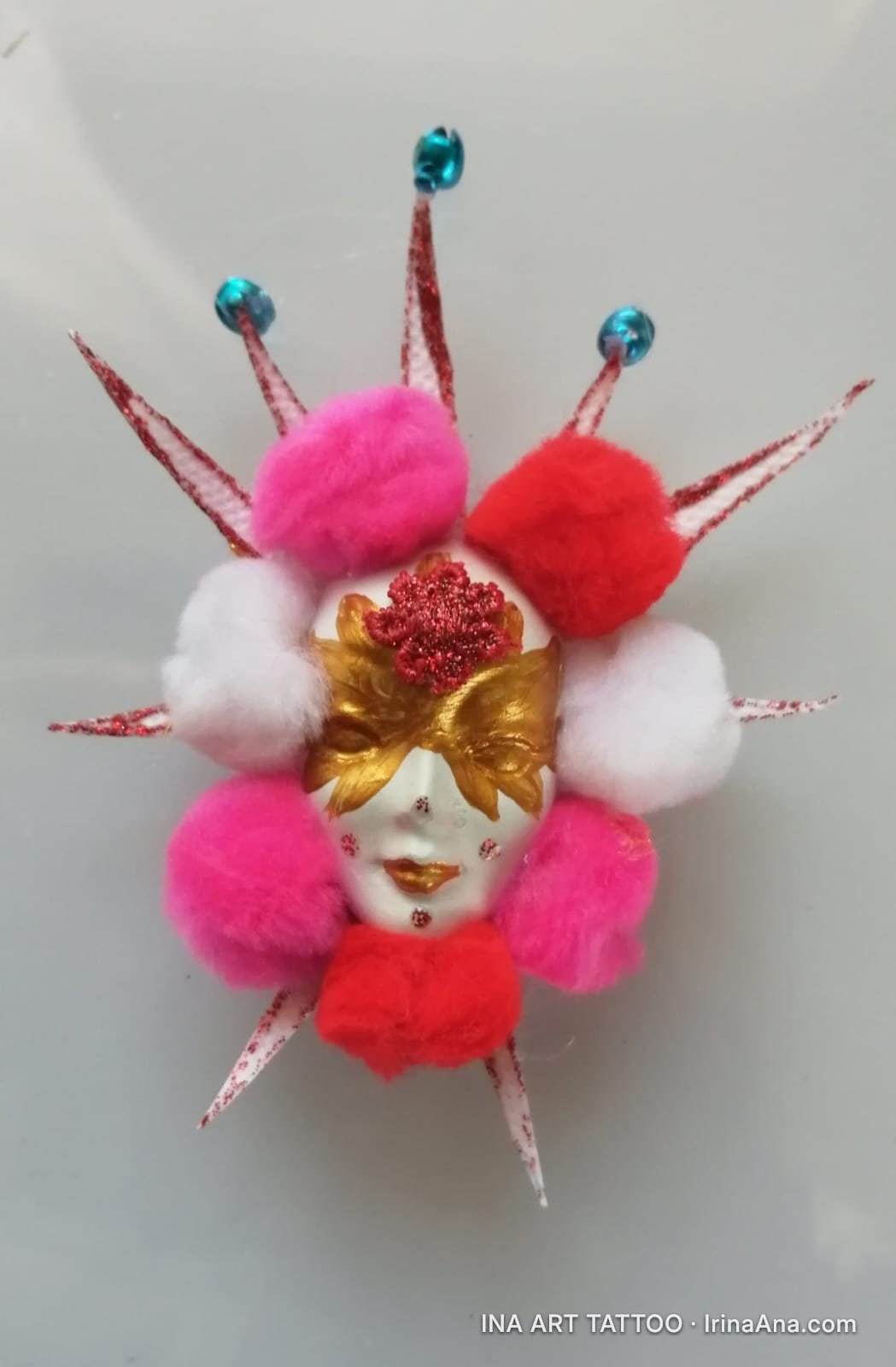 A white Venetian mask with golden eyes and red lips, decorated with fluffy pink, red, and white pom-poms, and glittery spike…
