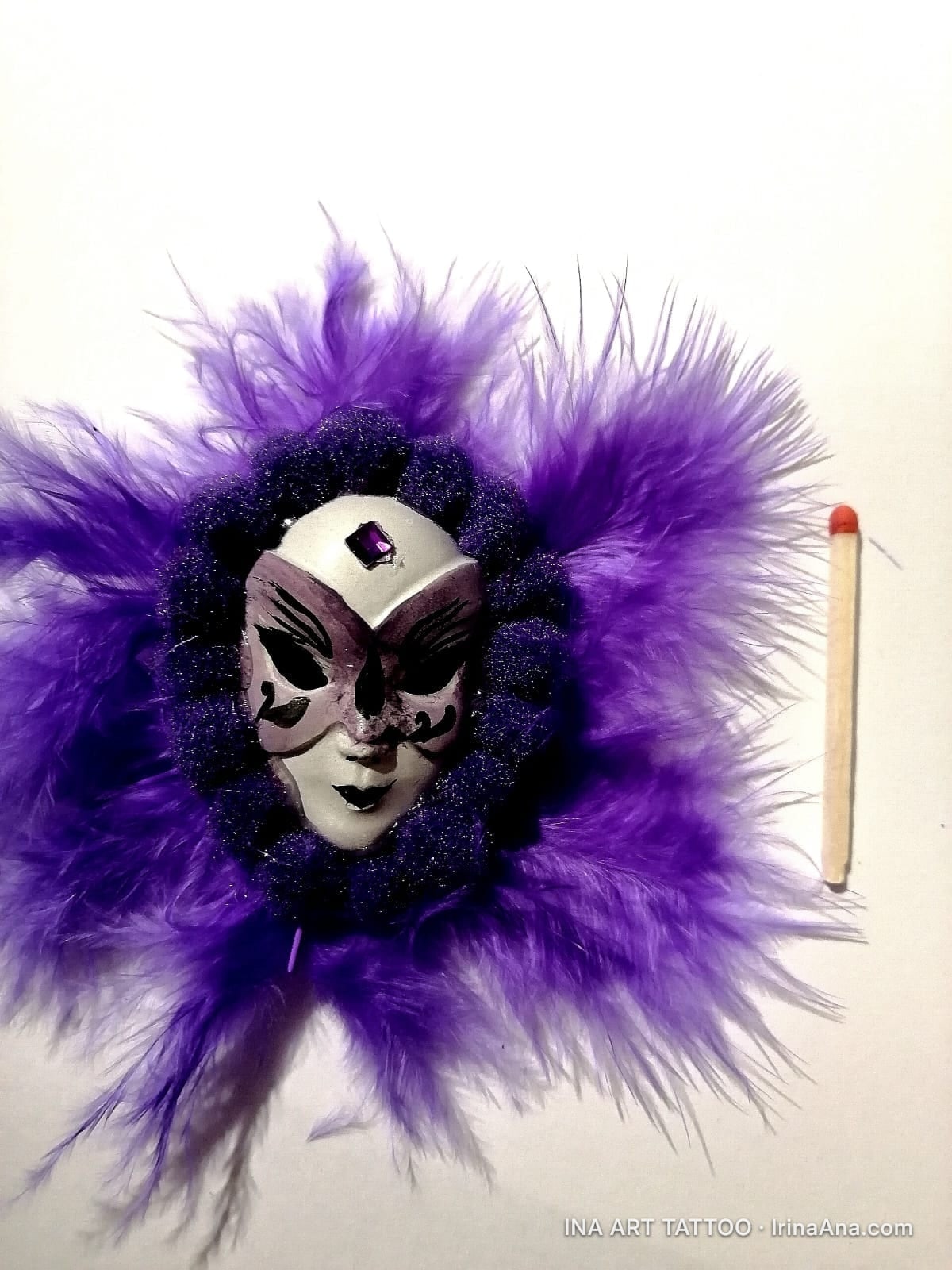 A mărțișor with a white and purple Venetian mask, surrounded by fluffy purple feathers. A matchstick is beside it for scale.