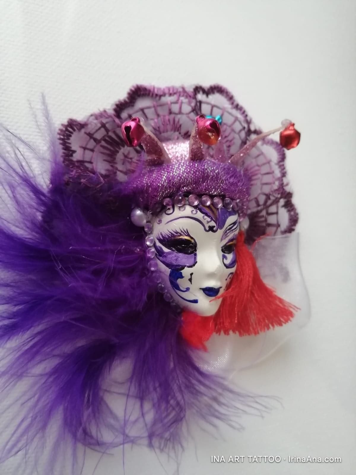 A white Venetian mask, decorated with purple and blue, with purple feathers and red tassels, on a white background.