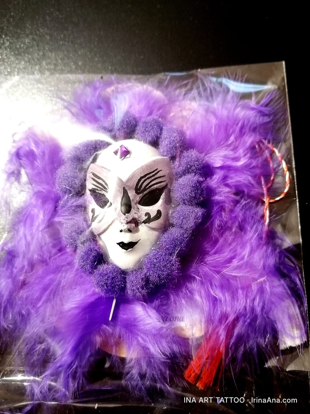 Mărțișor: a white Venetian mask with black and purple details, surrounded by purple feathers and pom-poms, with a red-white…