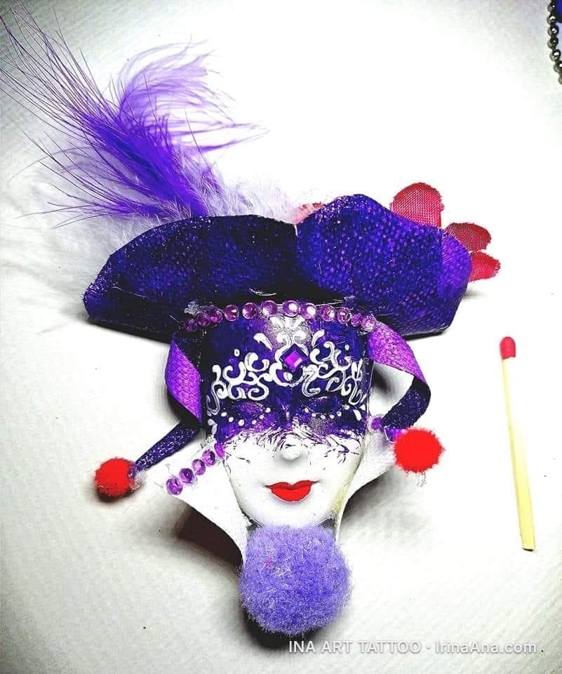 Mărțișor: white and purple Venetian mask with feathers, glitter, and red pompoms. A matchstick is next to it for scale.