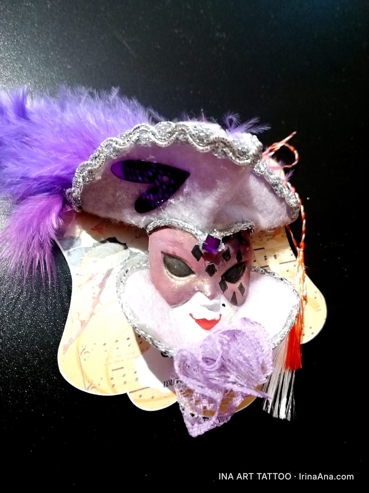 Mărțișor: decorative Venetian mask with purple feathers, white ruffle collar, and silver details, on a black background.