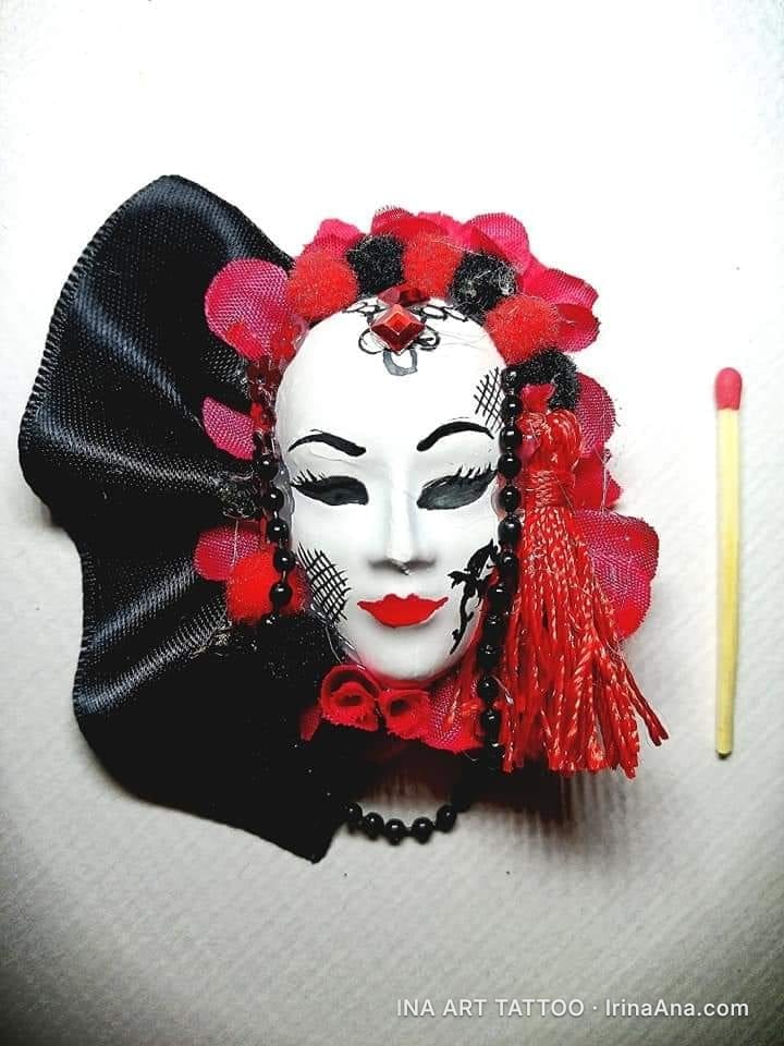 A white Venetian mask, decorated with red and black flowers, ribbons, and beads. A matchstick is next to it.