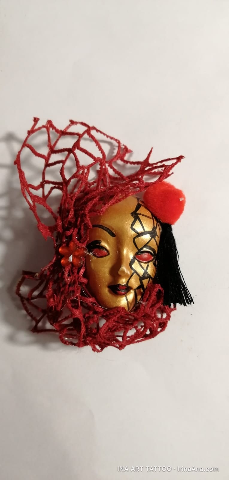 A golden mask with red eyes, surrounded by red threads and a red pompom, on a white background.