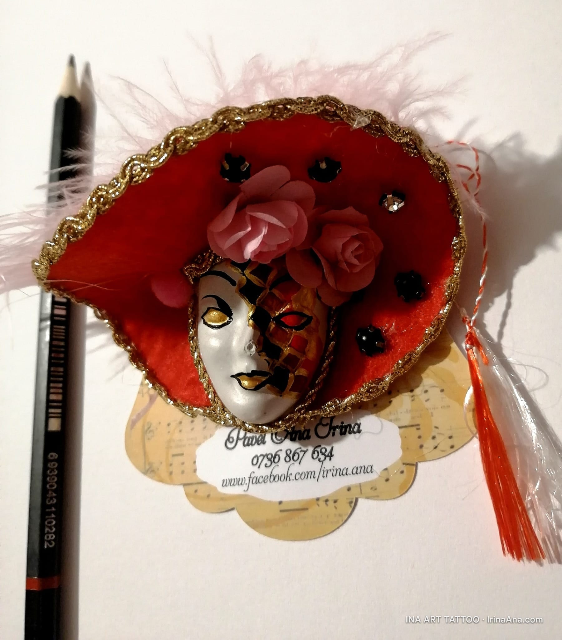 A handmade Mărțișor featuring a white and gold Venetian mask, set on a red hat decorated with flowers and feathers.