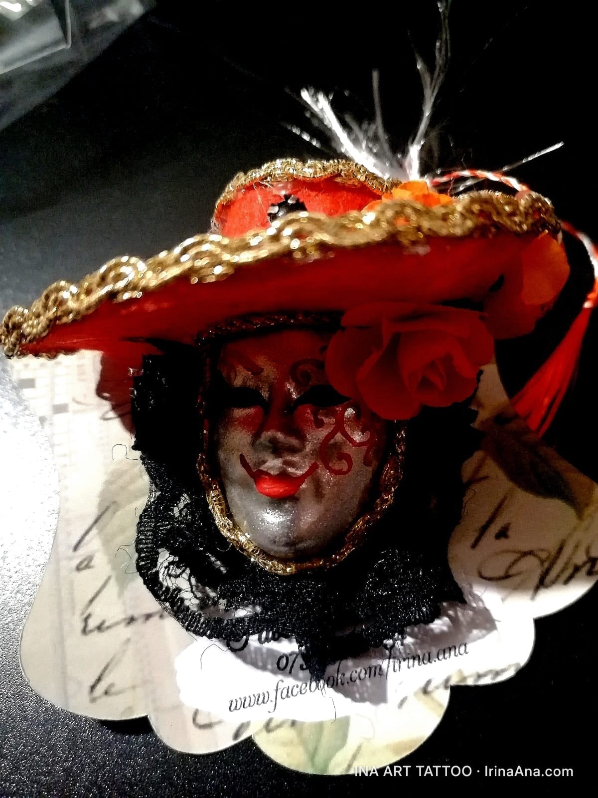 A handmade craft object shaped like a Venetian mask, featuring a red hat decorated with gold lace and white feathers.