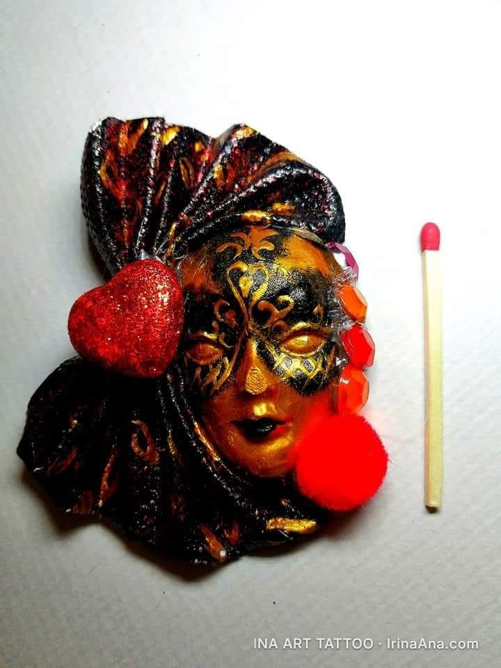 A decorated Venetian mask, predominantly in gold and black tones, with a glittery red heart and a red pompom, next to a matc…