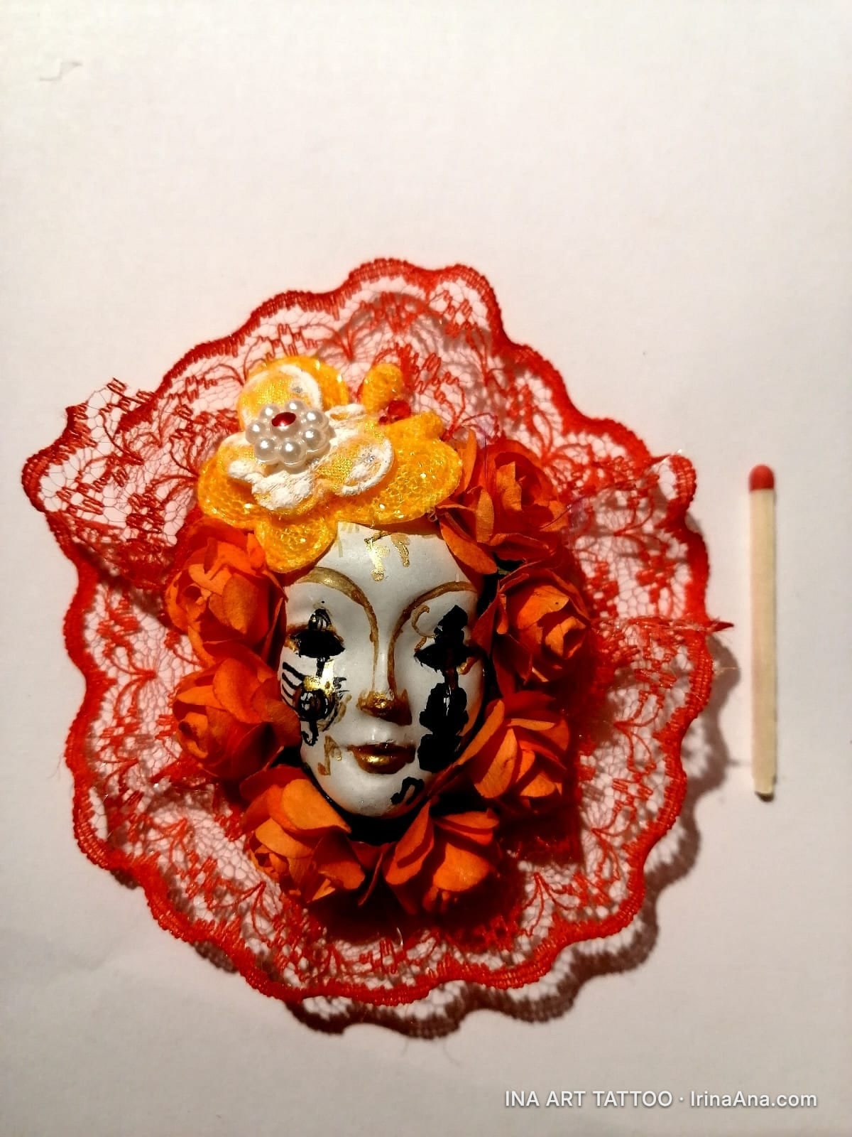 Handmade brooch featuring a white Venetian mask, surrounded by red lace and orange flowers, on a white background.