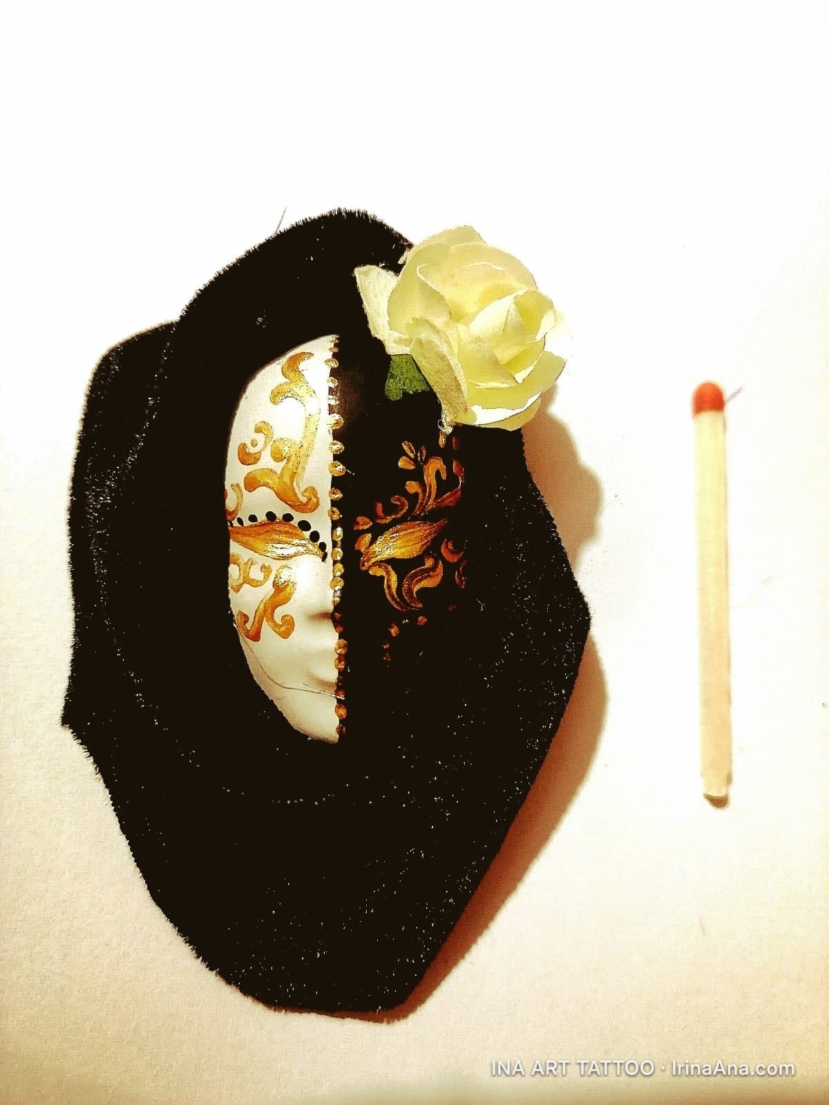 A black and white Venetian mask with gold details, resting on a piece of black fabric, with a white rose. A matchstick is ne…
