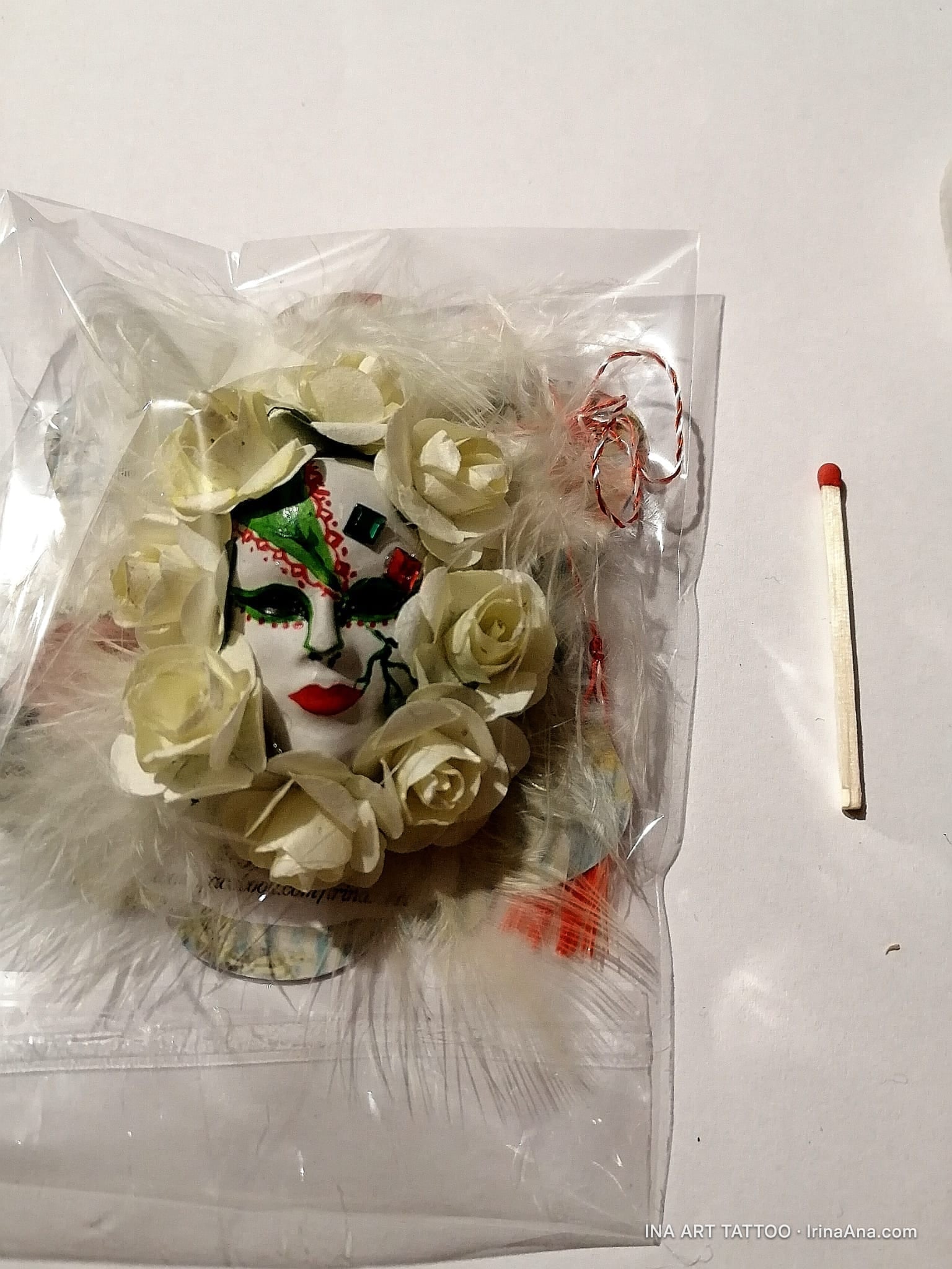 A white Venetian mask with red lips and green and red decorations, surrounded by white roses and feathers, inside a clear ba…