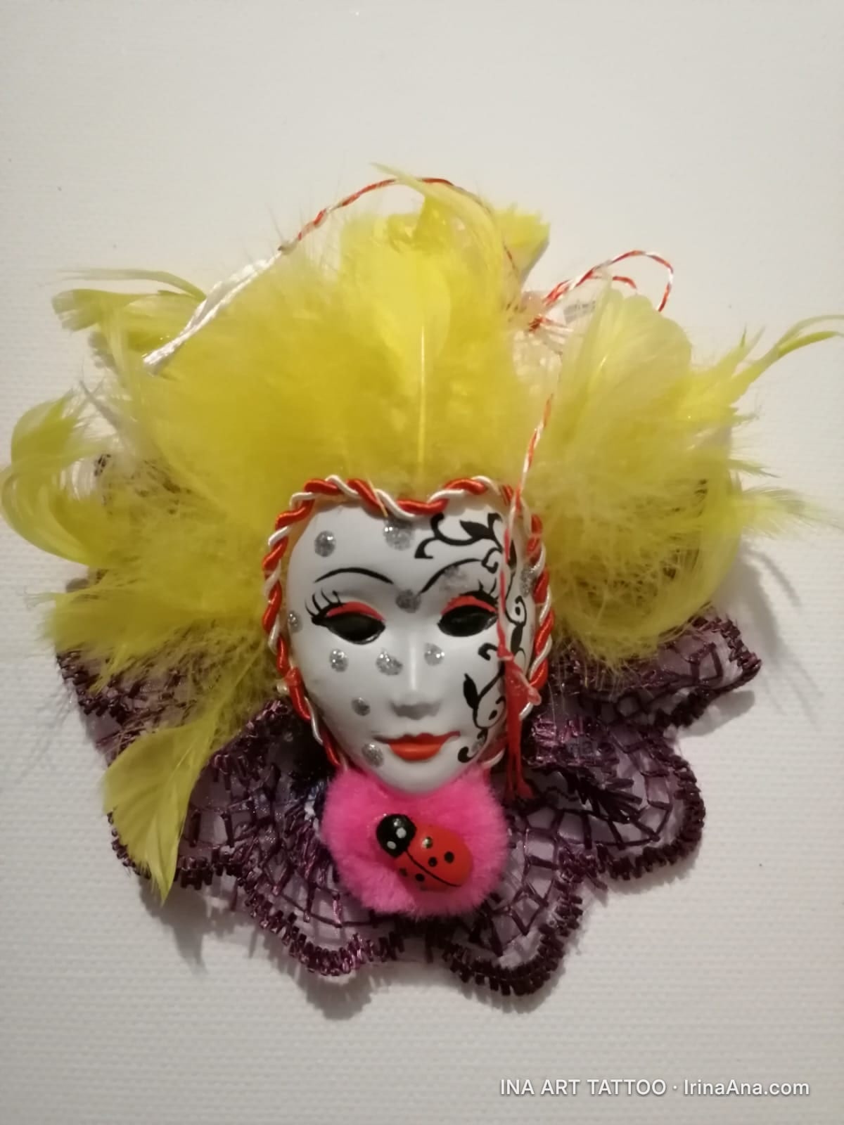Mărțișor: white Venetian mask with black pattern, surrounded by yellow feathers and purple lace, with a pink ladybug.