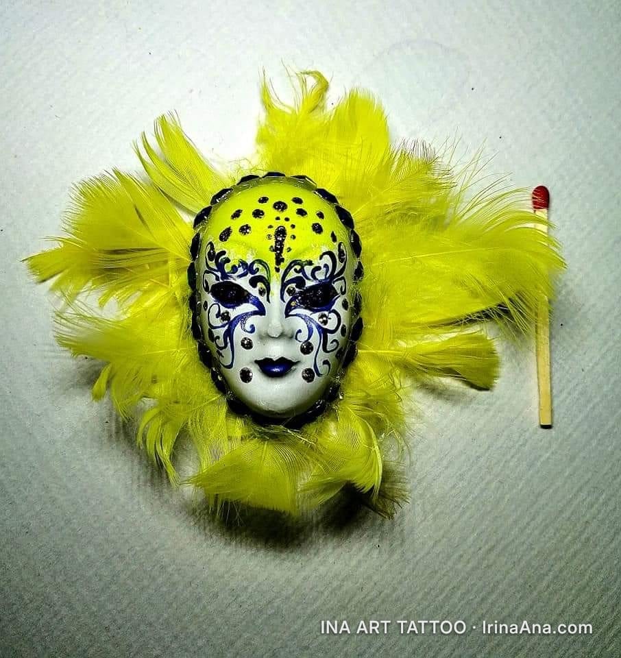 A white Venetian mask with blue and black details, surrounded by yellow feathers. A matchstick is next to it.