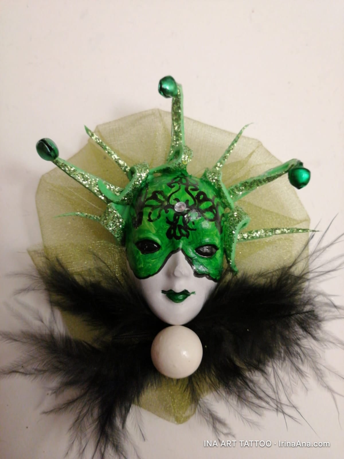 A harlequin-style carnival mask with a white face, green glitter mask with black details, black feathers, and a white ball.