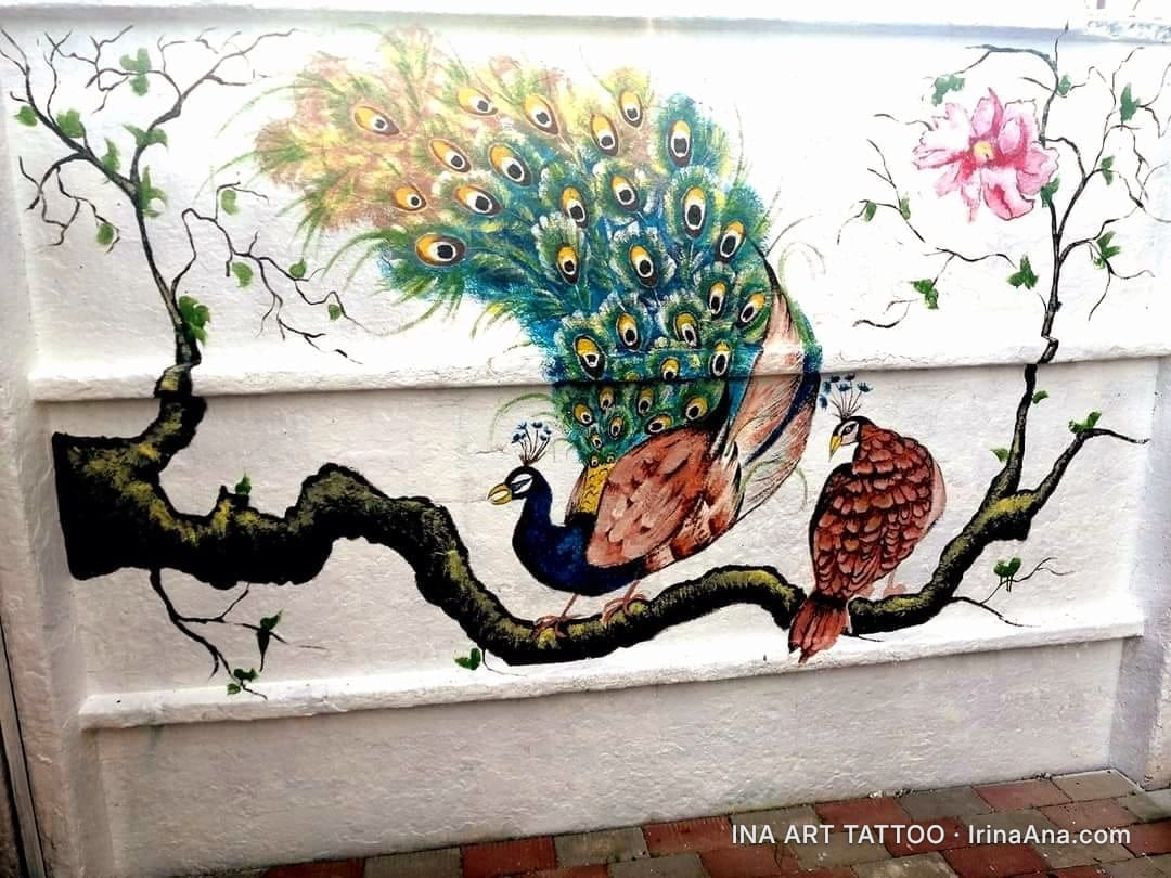 Pictură murală cu doi păuni pe o creangă groasă de copac, cu pene viu colorate și o floare roz în fundal.