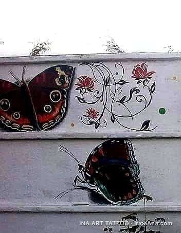 Doi fluturi maro-roșcați cu ochiuri pe aripi și un model floral cu flori roșii și frunze negre, pictate pe un perete.