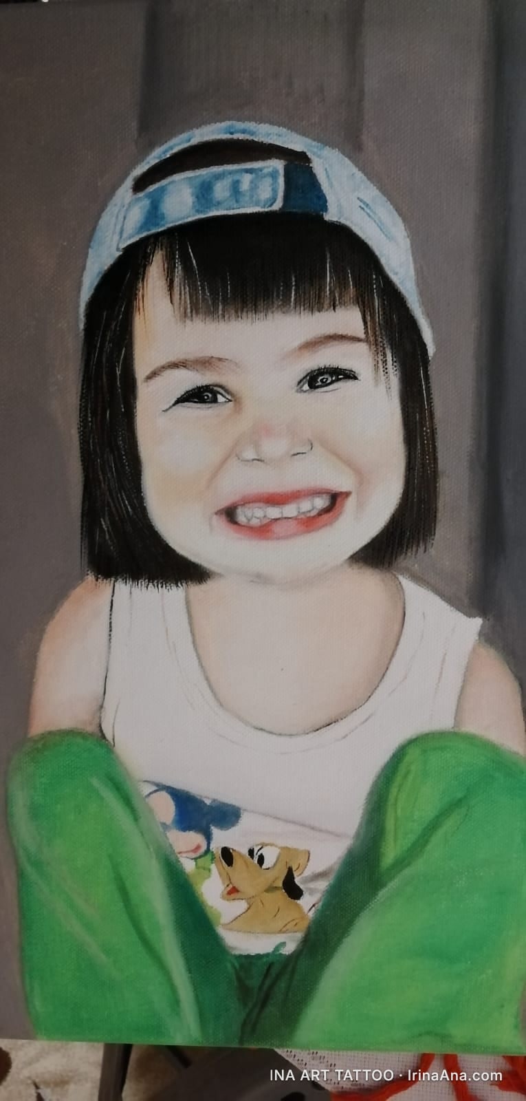 Portrait of a smiling child with short hair, bangs, and a blue cap, wearing a white tank top and green pants.