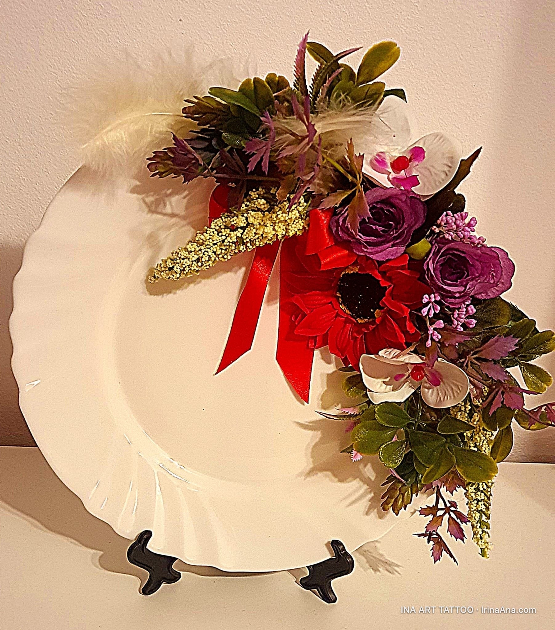 O farfurie decorativă albă, ondulată, cu un aranjament floral bogat, roșu și violet, cu o pană albă, pe partea superioară dr…