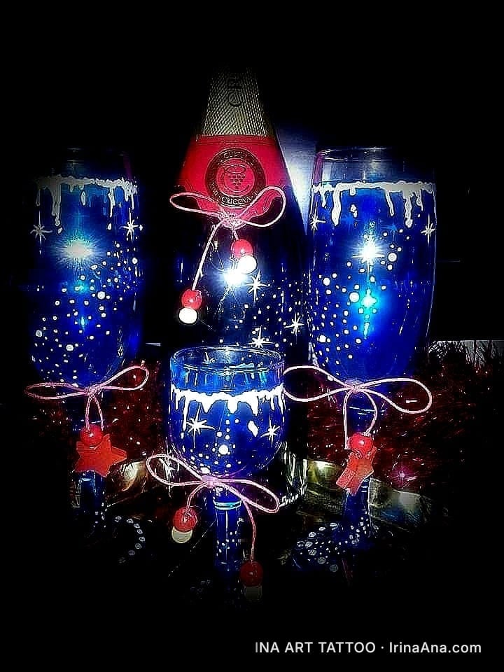 O sticlă de șampanie și trei pahare albastre, decorate cu fulgi de nea albi, steluțe și panglici roșii, pe fundal negru.