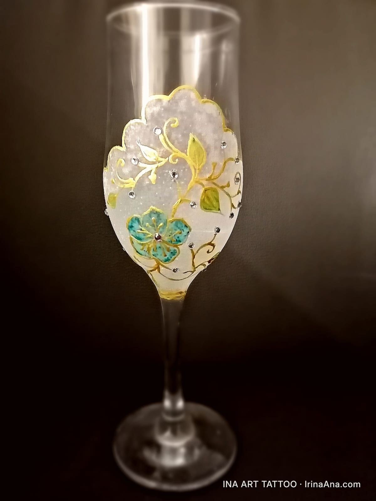Pahar de șampanie din sticlă, decorat cu un model floral alb, auriu și turcoaz, presărat cu mici pietre strălucitoare.