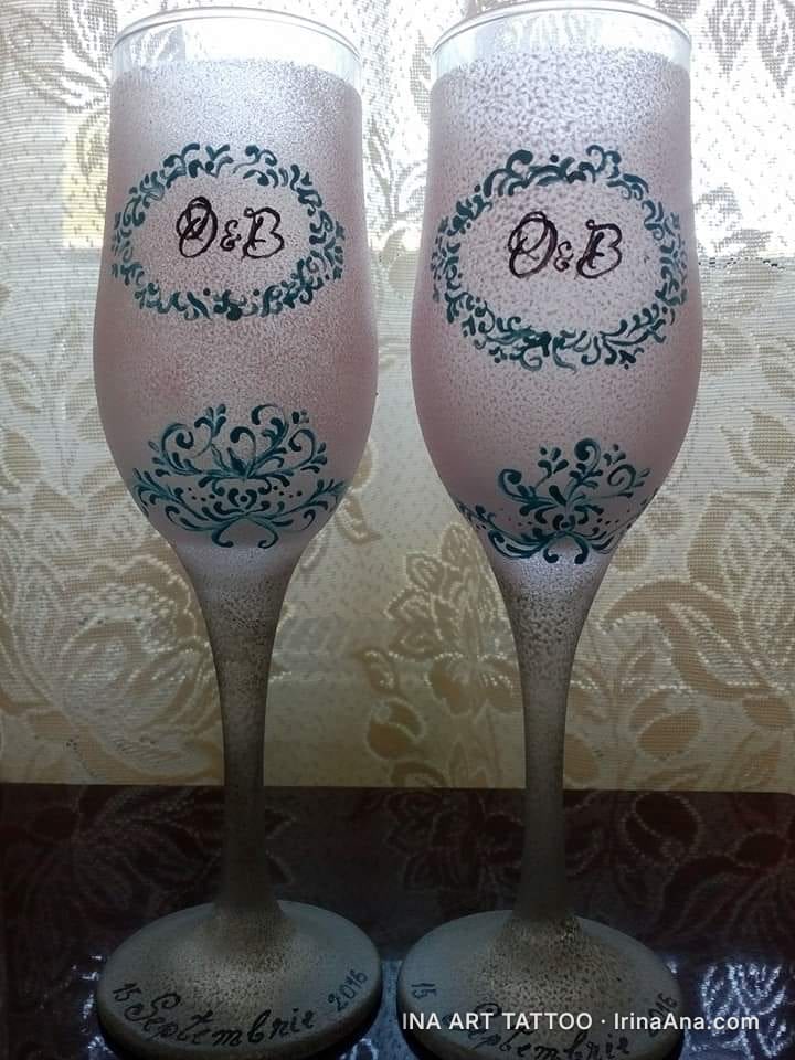 Două pahare de șampanie decorate cu inițialele D&B înconjurate de un chenar verde-albăstrui, pe un fundal rozaliu. Pe bază s…
