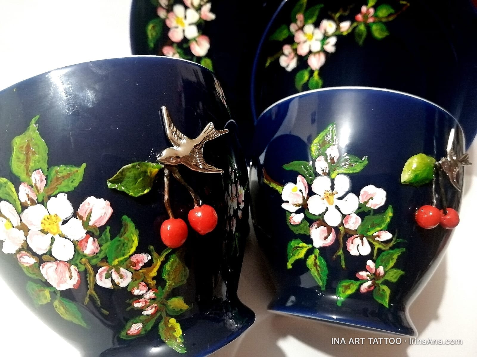 Două cești de ceramică albastru închis, decorate cu flori albe de cireș, frunze verzi și cireșe roșii 3D. O pasăre metalică…