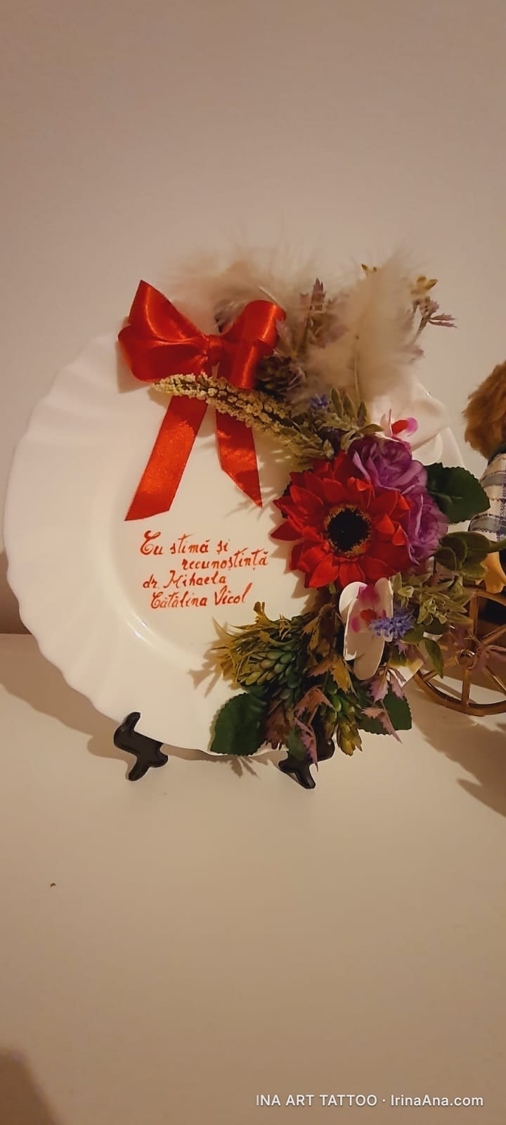 O farfurie decorativă albă, cu o fundă roșie și un aranjament floral colorat cu flori roșii, mov și frunze verzi.