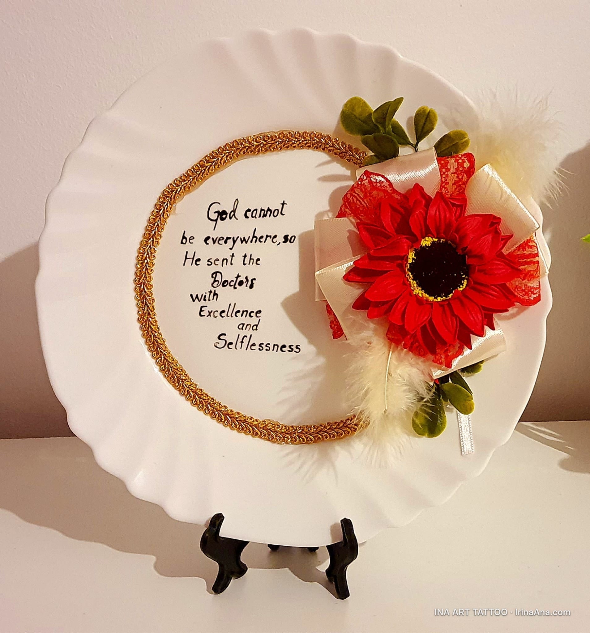 Farfurie decorativă albă cu o floare roșie din material textil, panglici, pene și un citat caligrafic.