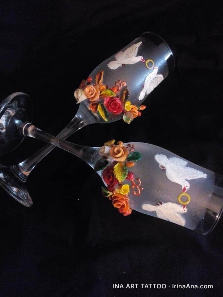 Două flûte de șampanie decorate cu porumbei albi și trandafiri miniaturali din lut polimeric, pe fundal negru.