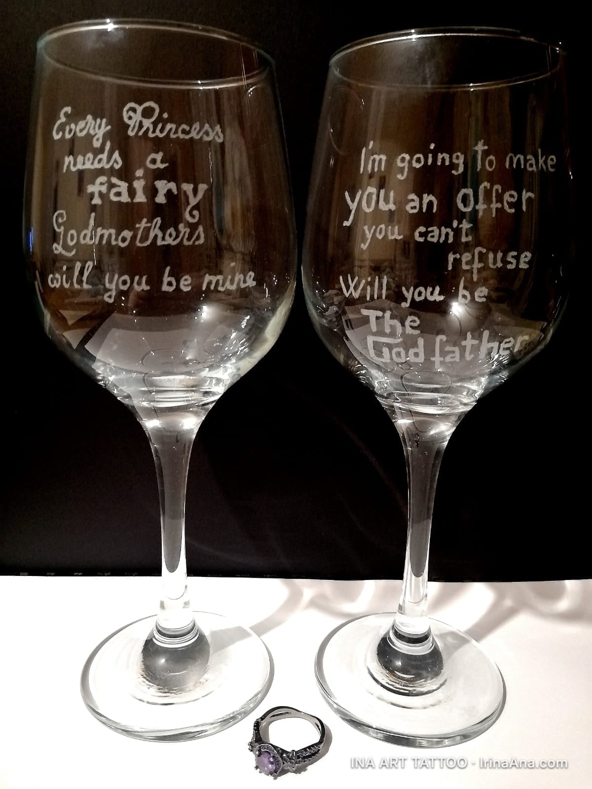Două pahare de vin din sticlă, gravate cu mesaje pentru nași, și un inel cu piatră mov la bază.