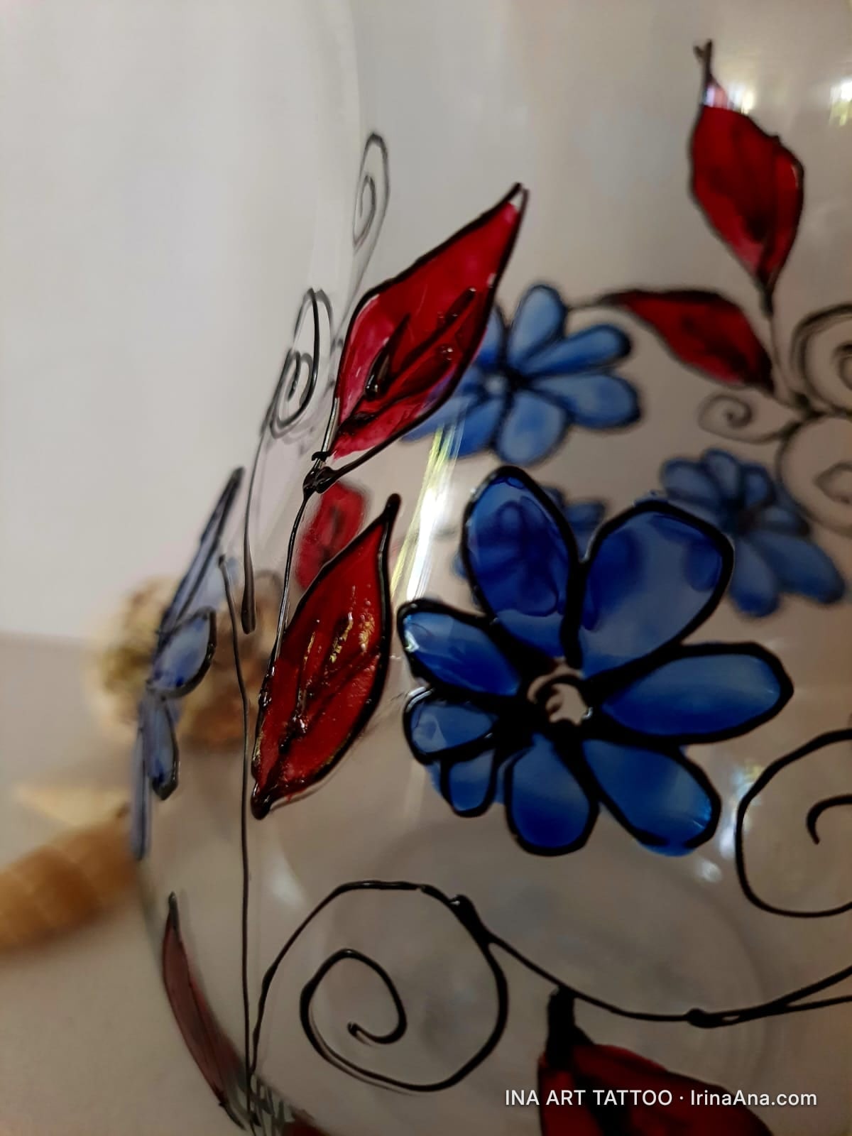 Vază din sticlă transparentă, decorată cu flori albastre și frunze roșii, conturate cu negru.