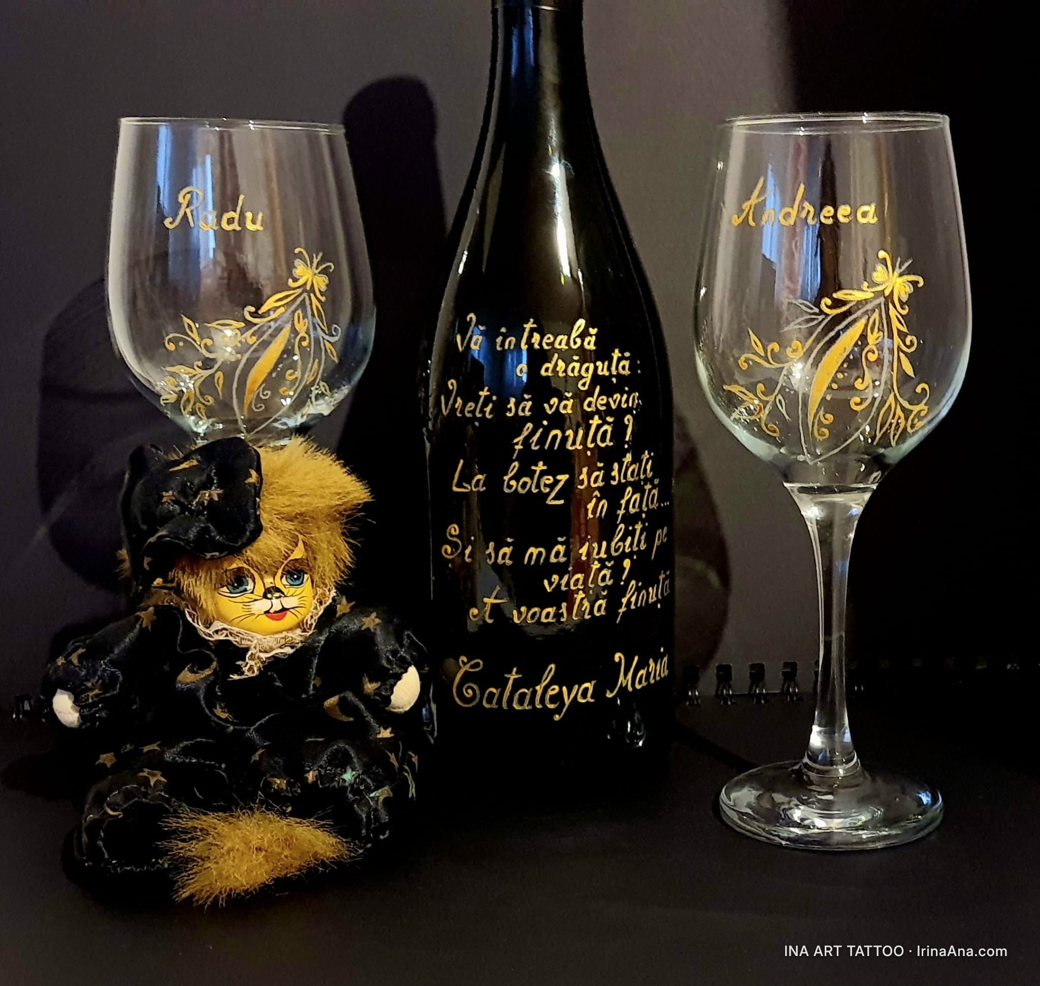 Două pahare de vin și o sticlă neagră, pictate manual cu auriu, lângă o păpușă pisică. Pe pahare sunt nume, pe sticlă un tex…