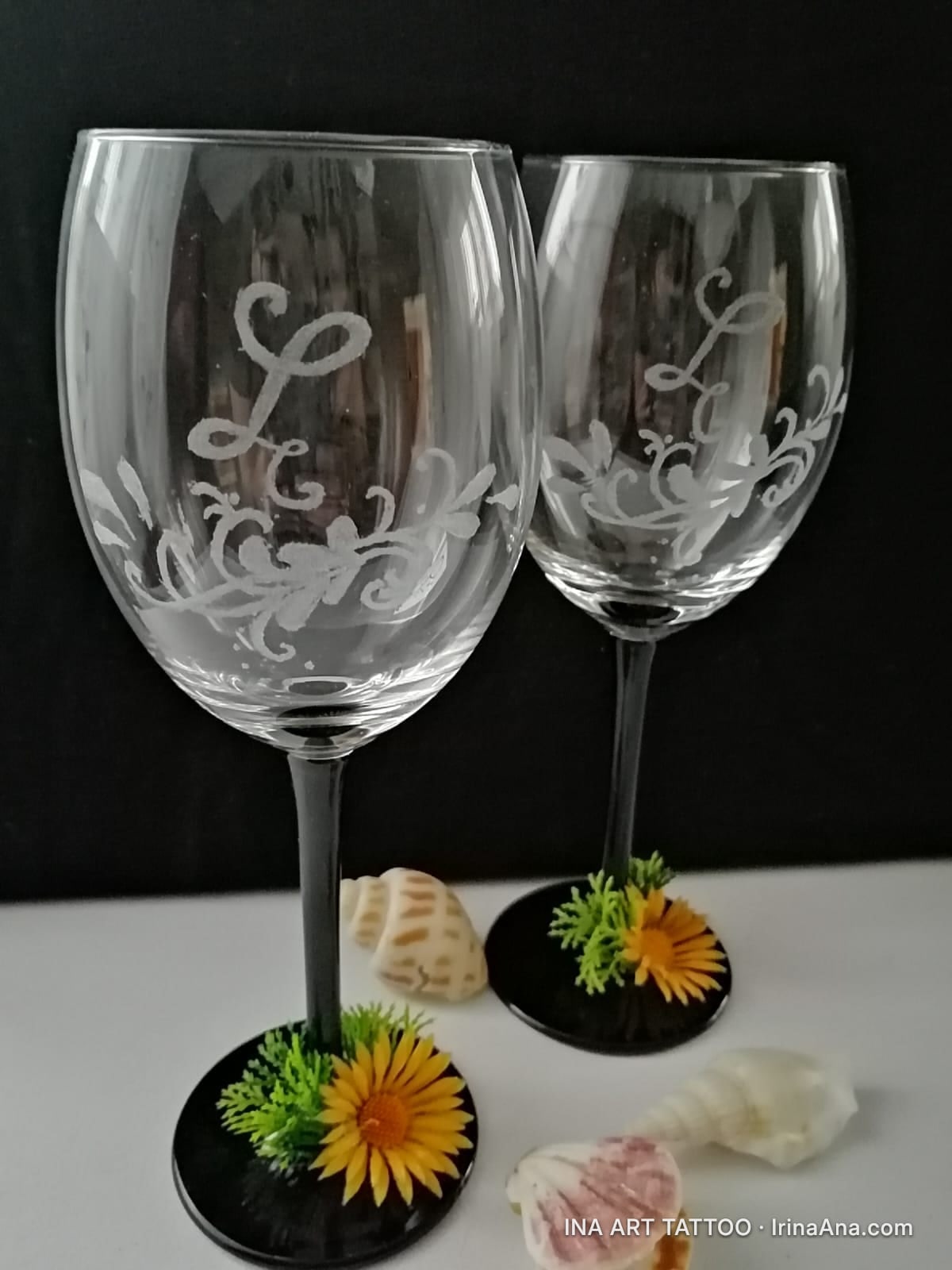 Două pahare de vin din sticlă, cu picior negru, decorate cu monograme L și flori portocalii la bază, lângă scoici.